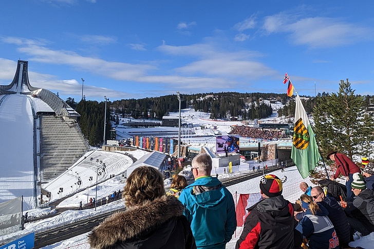 Zuschauergruppe auf einer Tribüne blickt auf eine Skisprungschanze und eine schneebedeckte Wettkampfanlage mit Zuschauermenge im Hintergrund.