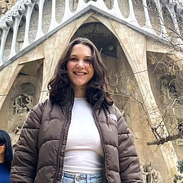 Woman wearing a brown puffer jacket and white t-shirt standing in front of a large stone facade with gothic arches and sculptures.
