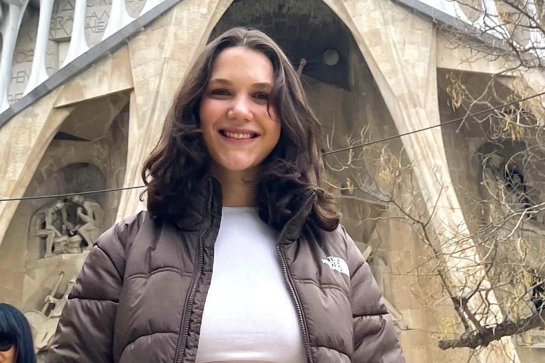 Frau mit brauner Steppjacke und weißem T-Shirt vor einer großen, steinernen Fassade mit gotischen Bögen und Skulpturen.