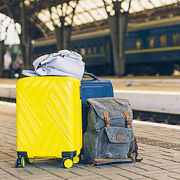 A yellow suitcase, a blue rucksack and a grey bag on a train station platform, with a blue train in the background.