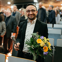 Dr. Haci Cevik trägt einen dunkelfarbenen Sakko, ein helles Hemd, hält einen Blumenstrauß mit gelben und orangen Blumen in der Hand und blickt freundlich in die Kamera. Im Hintergrund ist ein Hörsaal mit Besucher*innen der Preisverleihung zu sehen.