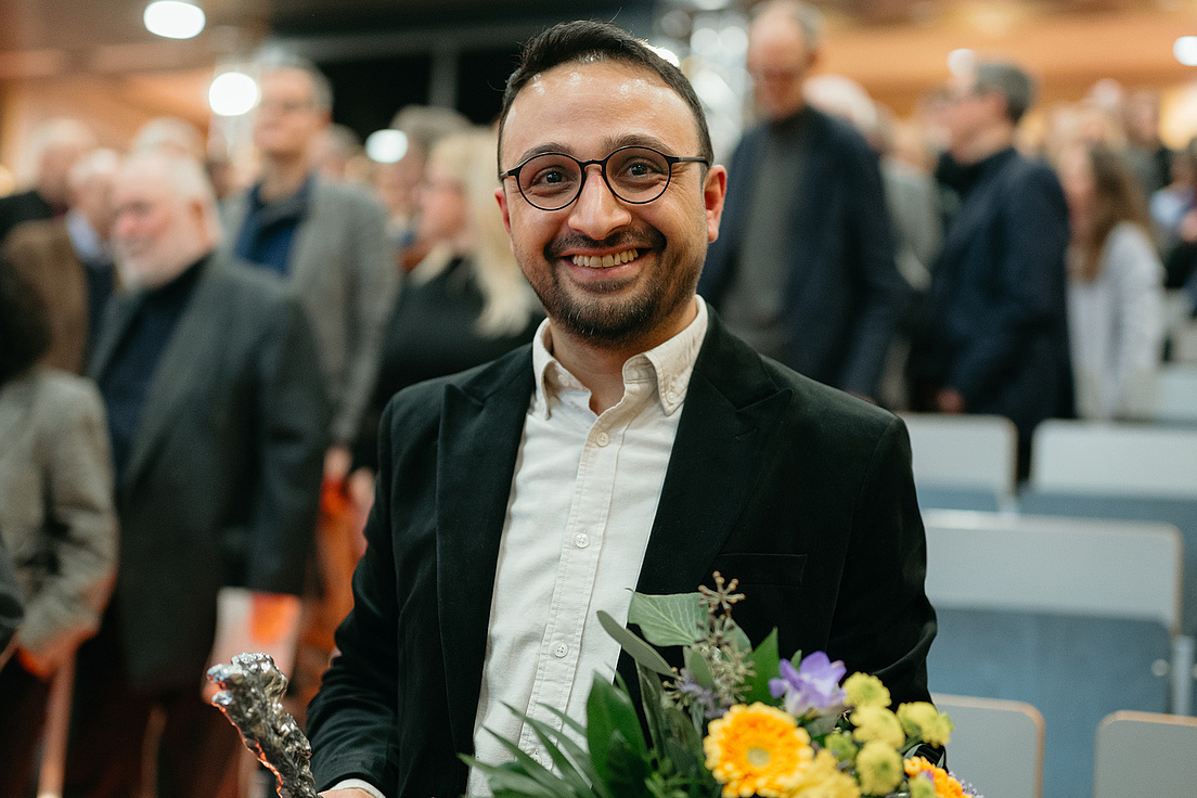 Dr. Haci Cevik trägt einen dunkelfarbenen Sakko, ein helles Hemd, hält einen Blumenstrauß mit gelben und orangen Blumen in der Hand und blickt freundlich in die Kamera. Im Hintergrund ist ein Hörsaal mit Besucher*innen der Preisverleihung zu sehen.