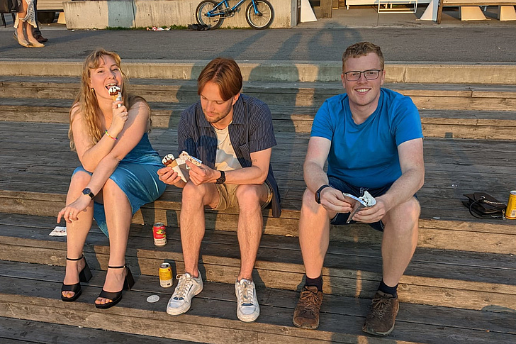 Drei Personen sitzen auf Holzstufen und essen Eis, zwei Dosen stehen auf den Stufen neben ihnen.