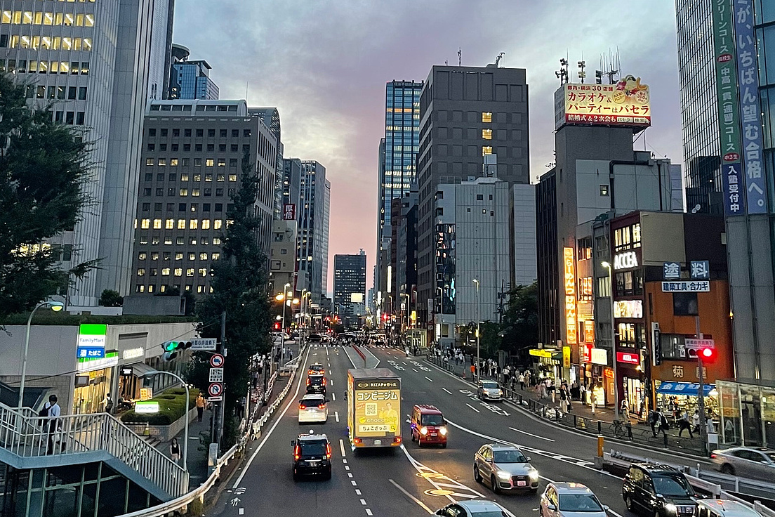 Straße in einer Stadt mit mehrspuriger Fahrbahn, umgeben von hohen Gebäuden und beleuchteten Werbetafeln bei Dämmerung