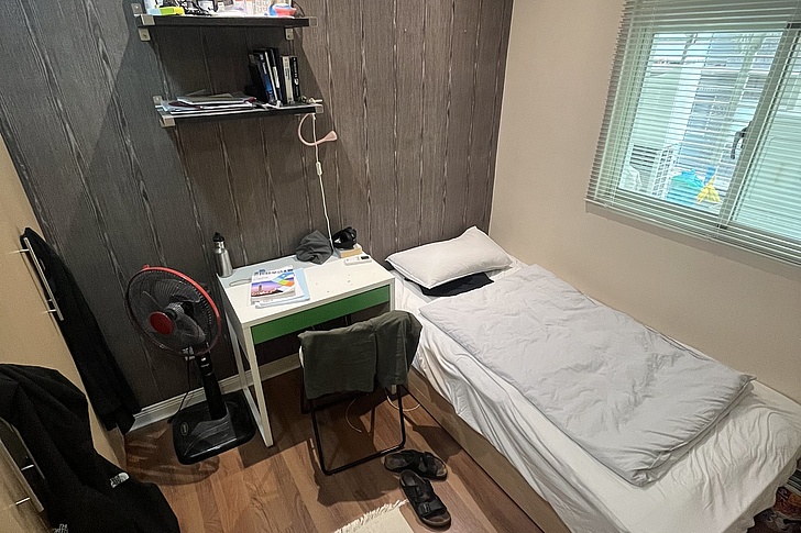Kleines Schlafzimmer mit Einzelbett, Schreibtisch, Stuhl, Ventilator und Fenster mit Jalousien
