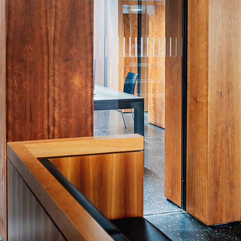 Grimm-Zentrum Das Bild zeigt einen modernen Innenraum mit einem klaren Fokus auf Holzelemente und Glas. Im Vordergrund ist ein Holzgeländer zu sehen, das glatt und poliert aussieht und eine warme, natürliche Farbe hat. Das Geländer verläuft diagonal durch das Bild und trennt den Vordergrund vom Hintergrund.