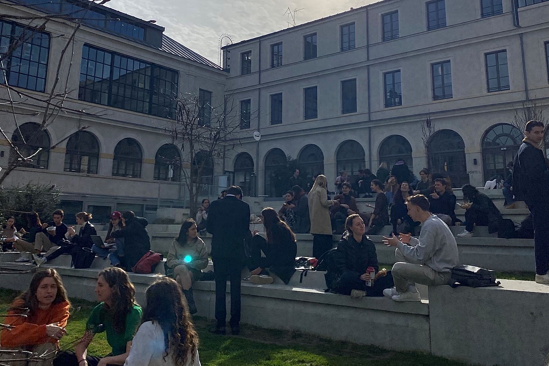 Menschen sitzen und stehen auf einer mehrstufigen Freitreppe vor einem großen Gebäude mit vielen Fenstern.