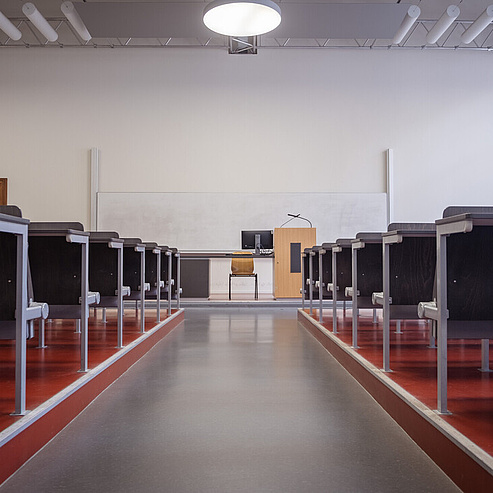 Das Bild zeigt einen leeren Hörsaal oder Vortragssaal. Der Raum ist symmetrisch eingerichtet mit Reihen von Sitzplätzen, die aus schwarzen Tischen und Stühlen bestehen. Die Tische sind an den Seiten des Raumes angeordnet, wobei ein breiter Mittelgang in der Mitte führt. Am Ende des Raumes steht ein Podium mit einem Mikrofon und einem Monitor darauf. An der Wand hinter dem Podium befindet sich eine große Tafel.