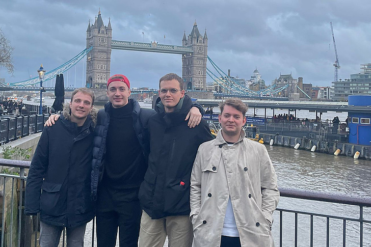 Vier Personen stehen an einem Geländer mit Blick auf die Tower Bridge und die Themse in London.