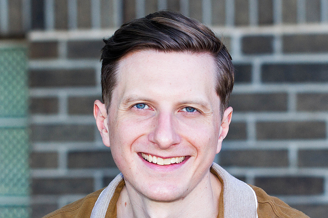 Torben Ott mit kurzen, dunkelbraunen Haaren und blauen Augen lächelt freundlich in die Kamera. Er trägt ein braunes Polohemd mit einem hellen, gestreiften Kragen. Der Hintergrund besteht aus einer grauen Ziegelwand.