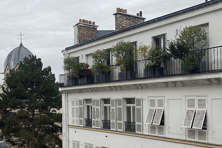 Mehrstöckiges weißes Wohnhaus mit Balkonen und geöffneten Fensterläden vor bewölktem Himmel und Baum im Vordergrund