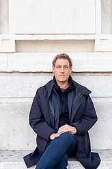 Joerg Fingerhut sitzt mit übergeschlagenen Bein vor einem Marmordenkmal und trägt eine dunkle Jacke.