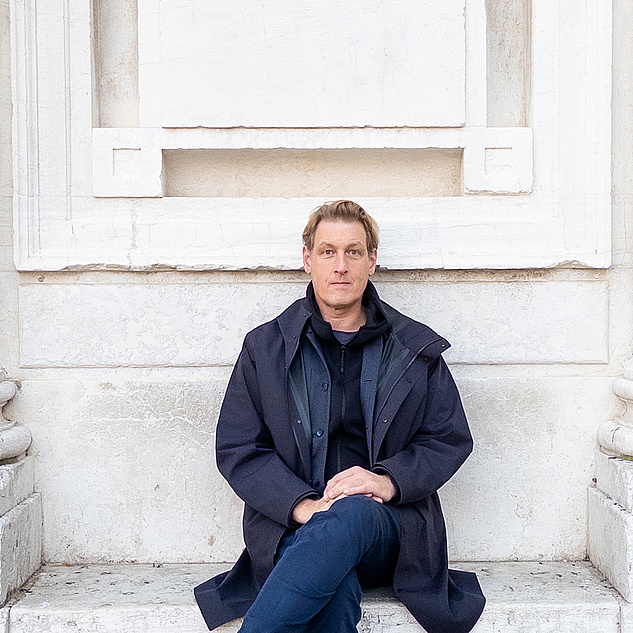 Joerg Fingerhut Joerg Fingerhut sitzt mit übergeschlagenen Bein vor einem Marmordenkmal und trägt eine dunkle Jacke.