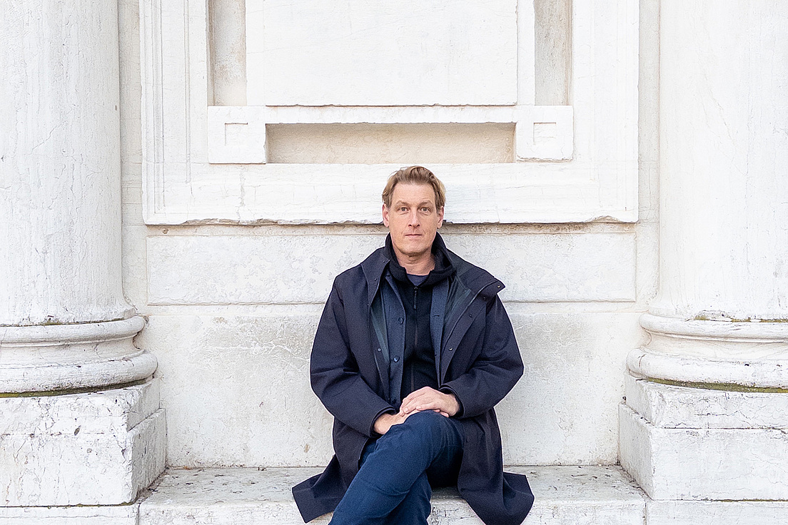 Joerg Fingerhut sitzt mit übergeschlagenen Bein vor einem Marmordenkmal und trägt eine dunkle Jacke.