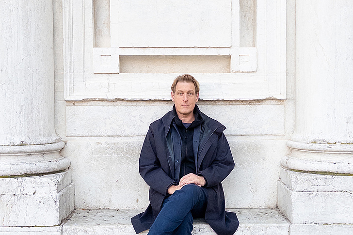 Joerg Fingerhut sitzt mit übergeschlagenen Bein vor einem Marmordenkmal und trägt eine dunkle Jacke.