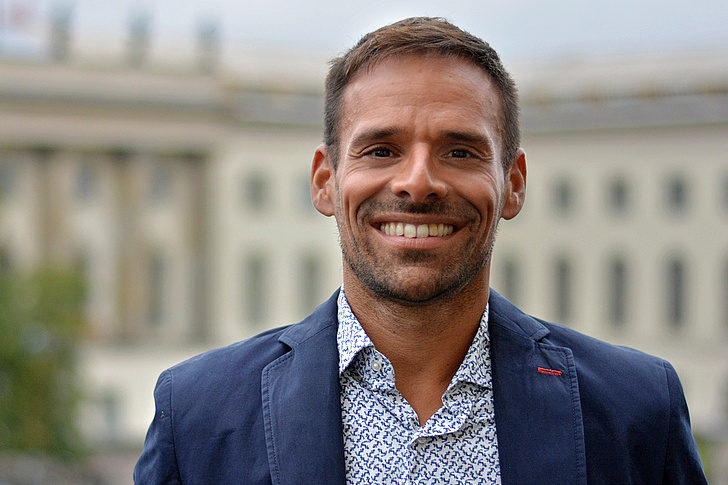 Yoan Vilain ist im Freien und lächelt in die Kamera und trägt ein kornblaues Hemd und ein blau-weiß gemustertes Hemd. Im Hintergrund ist das HU-Hauptgebäude zu sehen.