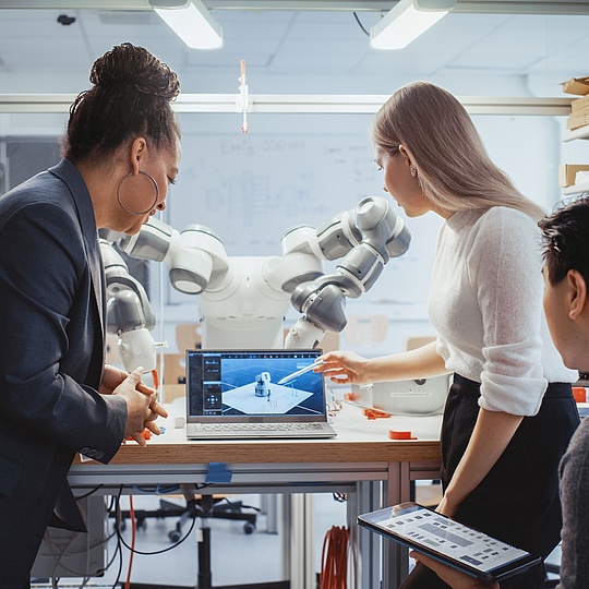 Diverse Team: Chief Engineer and Promising Computer Scientists Analyse and Discuss Optimisation of Autonomous Manufacturing Facility That Creates Great Products. Laboratory with Robotic Arm. Drei Personen arbeiten gemeinsam in einem modernen Labor oder Forschungsraum. Sie stehen um einen Tisch mit einem Laptop, auf dessen Bildschirm ein 3D-Modell eines Roboters zu sehen ist. Hinter ihnen befindet sich ein großer Industrieroboterarm.