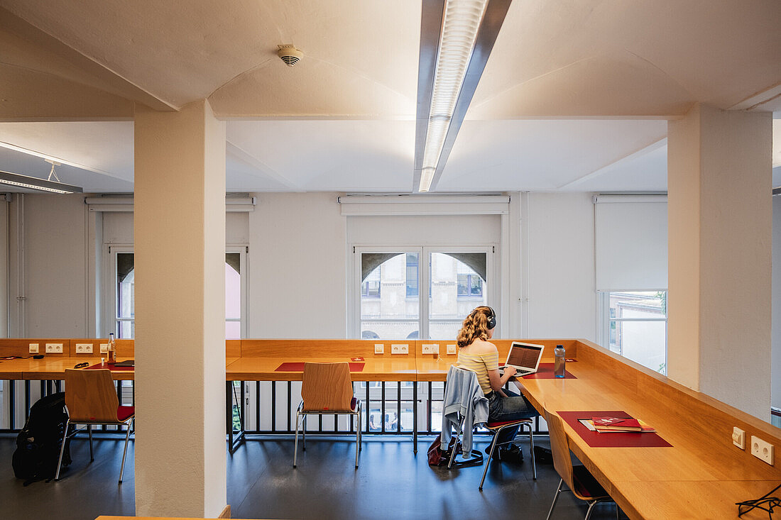 Eine Frau sitzt mit dem Rücken zur Kamera an einem Tisch. Sie hat einen Laptop vor sich und arbeitet an einem Arbeitsplatz. Man sieht lange Arbeitsplatten aus hellem Holz, vorne hohe helle Fenster, die viel Licht in den Raum lassen.