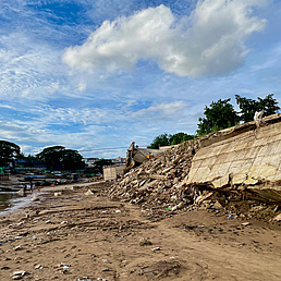 Zu sehen ist eine zerstörte Grenzinfrastruktur nach einem Monsun. Im Hintergrund räumen Personen vor Hausbooten im Flussbett auf 