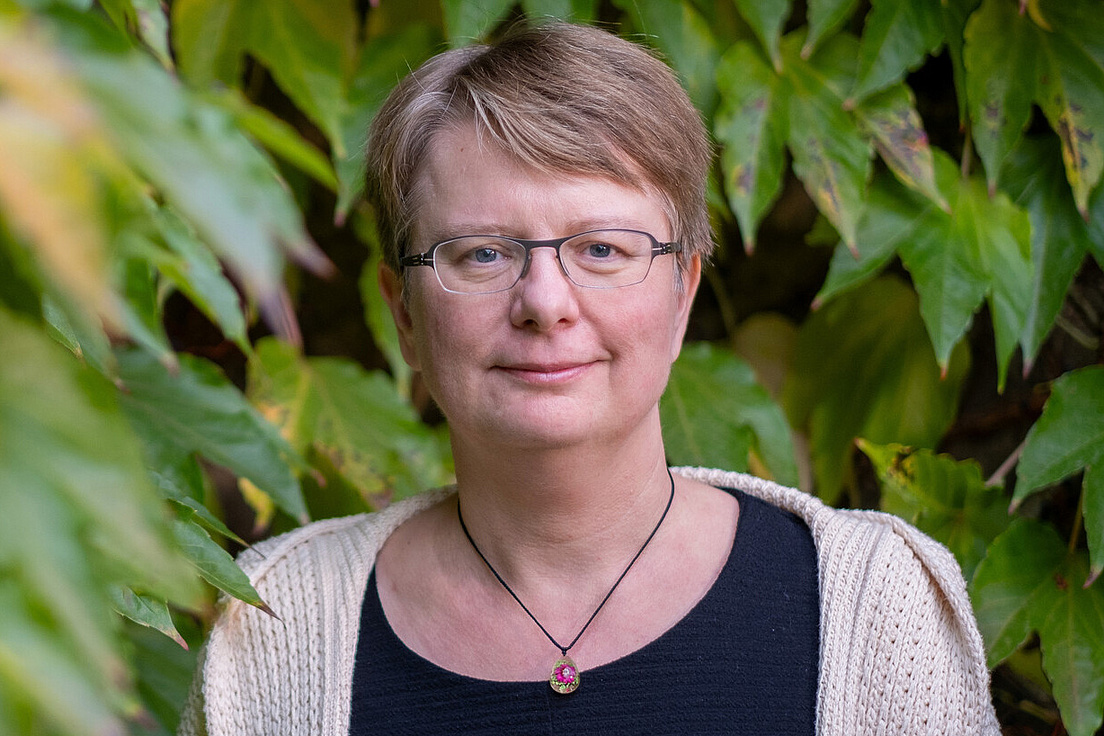 Kerstin Kaufmann trägt eine Kette um den Hals, ein schwarzes Shirt und eine beige Strickjacke. Im Hintergrund sind Blätter eines Baums zu sehen.