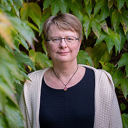 Kerstin Kaufmann trägt eine Kette um den Hals, ein schwarzes Shirt und eine beige Strickjacke. Im Hintergrund sind Blätter eines Baums zu sehen.