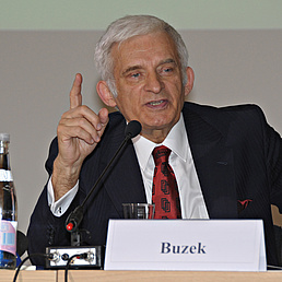 Jerzy Buzek sitzt mit erhobenem Finger an einem Tisch und beantwortet eine Frage aus dem Publikum. Vor ihm befinden sich Wasserflaschen und ein Namensschild.