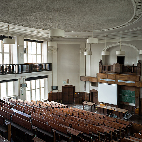 Das Bild zeigt einen großen, leeren Hörsaal in einem Gebäude mit historischem Flair. Der Raum ist mit Holzbänken und -tischen ausgestattet, die in Reihen angeordnet sind und zum Podium hinunterführen. An der Vorderseite des Raumes befindet sich ein Podium mit einem Pult und einer Tafel, auf der mathematische Formeln zu sehen sind. Über dem Podium hängt eine große Leinwand für Präsentationen.
