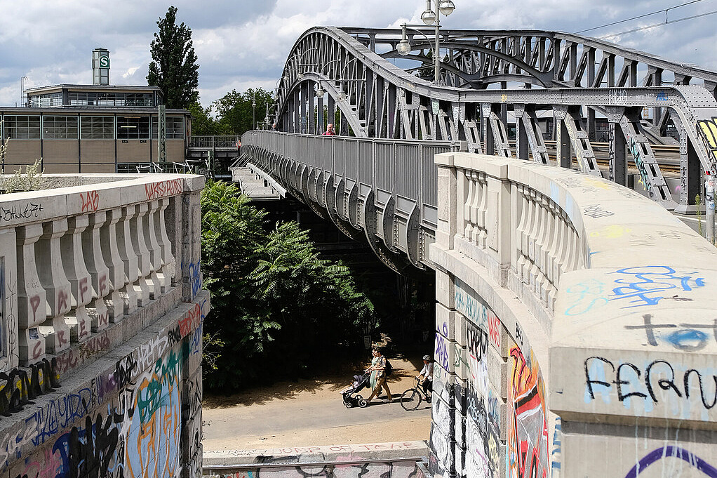 Bornholmer Brücke in Berlin übersät mit Graffiti. Untern läuft eine Familie mit Kinderwagen entlang, oben ist die Brücke leer bis auf einen Menschen, der sie überquert