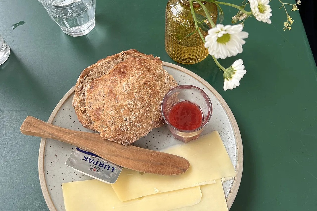 Teller mit zwei Scheiben Vollkornbrot, mehreren Scheiben Käse, einem kleinen Glas mit roter Marmelade, einem Holzmesser und einem kleinen Butterpäckchen auf grünem Tisch neben einer Vase mit weißen Blumen