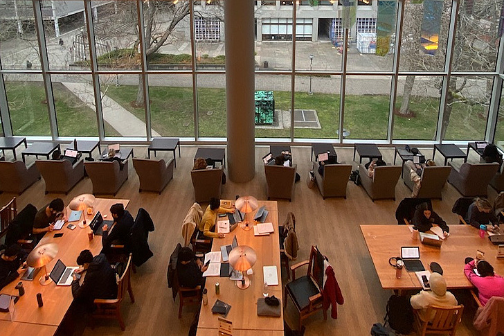Luftaufnahme einer belebten Universitätsbibliothek mit Studierenden an Holztischen mit Laptops, umgeben von bodentiefen Fenstern mit Blick auf Rasen, Bäume und Campuswege bei sonnigem Wetter.