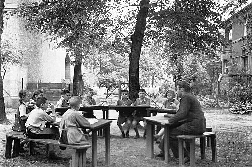 Max Bayer mit seinen Schülern beim Unterricht Diese Schwarz-Weiß-Fotografie zeigt eine Gruppe von Kindern und einen Erwachsenen, die im Freien an Holztischen und -bänken sitzen. Die Szene spielt sich auf einer Wiese oder einem Hof vor einem großen Baum ab, im Hintergrund sind Gebäude und weitere Bäume zu erkennen. Die Kinder, die meistens Hemden tragen, sitzen um die Tische herum und scheinen einer Erzählung oder einer Lektüre zu folgen, die der Erwachsene, der eine dunkle Jacke trägt, ihnen vorträgt.