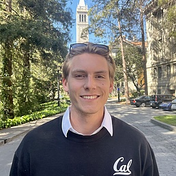 Young man wearing a black sweater with a white collar and 'Cal' logo standing on a tree-lined path with historic buildings in the background.