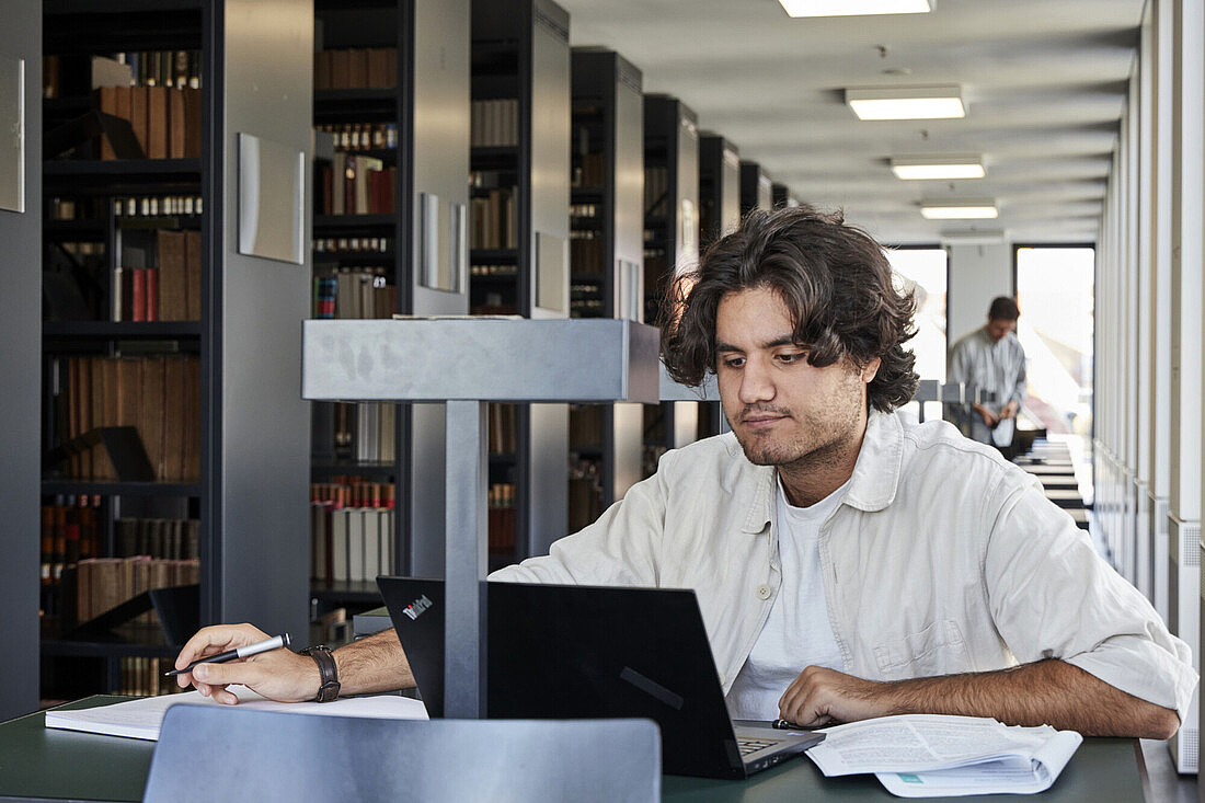 Ein junger Mann sitzt an einem Tisch in einer Bibliothek und arbeitet konzentriert an seinem Laptop. Er hält einen Stift in der Hand und hat Papiere vor sich liegen. Im Hintergrund sind Bücherregale und ein weiterer Mensch zu sehen, der ebenfalls beschäftigt ist. Die Bibliothek wirkt ruhig und gut beleuchtet.