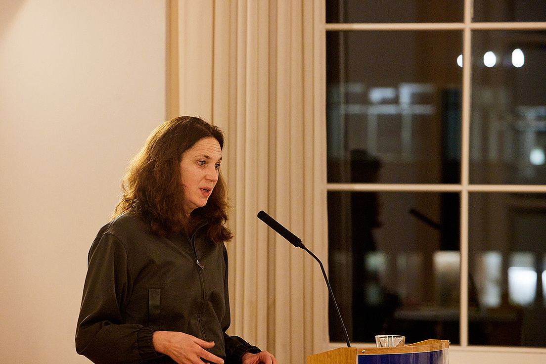 Viktoria Tkaczyk steht hinter einem Rednerpult und spricht ins Mikrofon anlässlich der Verleihung des Caroline von Humboldt-Preises. Sie trägt einen dunkelfarbenen Overall.