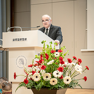 George Vella steht dem Publikum zugewandt am Rednerpult und hält seine Rede. Im Vordergrund befindet sich ein Blumengesteck.