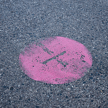 Das Bild zeigt einen rosa Kreis, der auf einer asphaltierten Oberfläche mit einem schwarzen Pinselstrich markiert ist. Der Hintergrund besteht aus grauem Asphalt mit kleinen Steinen und Kieseln.