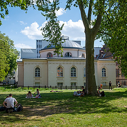 Campus Nord Ein kleines Parkgelände mit Blick auf das Tieranatomische Theater. Auf der Wiese sitzen Studierende.
