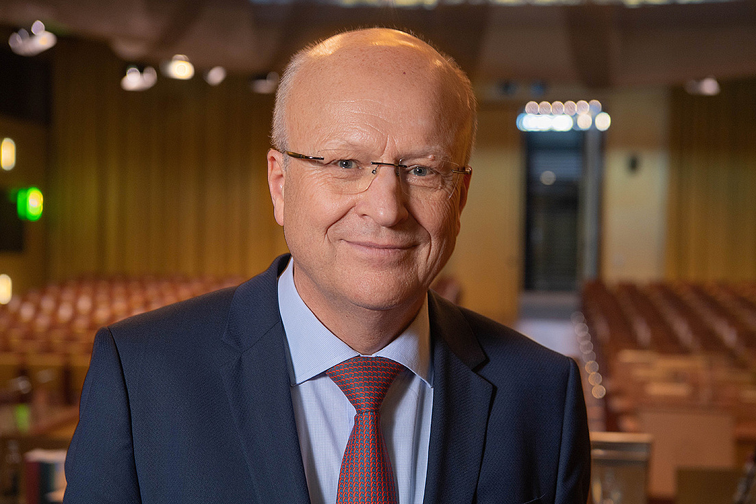 Koen Lenaerts trägt einen dunkelblauen Anzug, mit einem hellblauen Hemd und bordeaux-farbener Krawatte. Er trägt eine Brille und sieht freundlich in die Kamera. Im Hintergrund sind Stühle und Sitzreihen zu sehen.