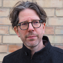 Ein Mann mit Brille und grauem Haar steht vor einer Ziegelwand. Er trägt einen dunklen Mantel.