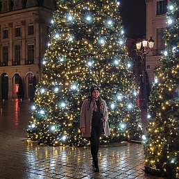 Frau mit Mütze in hellem Mantel steht abends zwischen zwei großen beleuchteten Weihnachtsbäumen mit gelben Lichtern auf dem festlich illuminierten Place Vendôme in Paris, umgeben von Arkaden und Laternen.