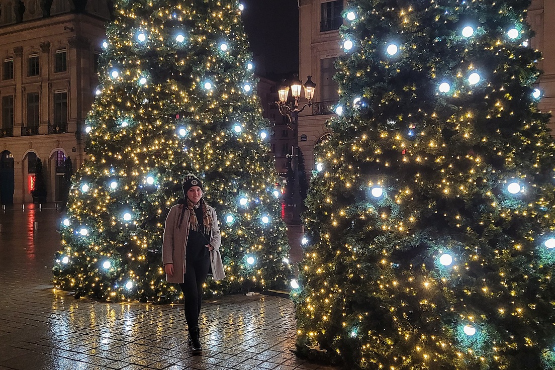 Frau mit Mütze in hellem Mantel steht abends zwischen zwei großen beleuchteten Weihnachtsbäumen mit gelben Lichtern auf dem festlich illuminierten Place Vendôme in Paris, umgeben von Arkaden und Laternen.