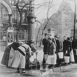 Menschen stehen an einem Brunnen und füllen Eimer mit Wasser, im Hintergrund Gebäude und Bäume.