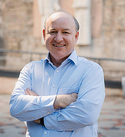 Matthew Larkum lächelt mit verschränkten Armen in die Kamera. Er trägt ein hellblaues Hemd und steht draußen vor einem Gebäude