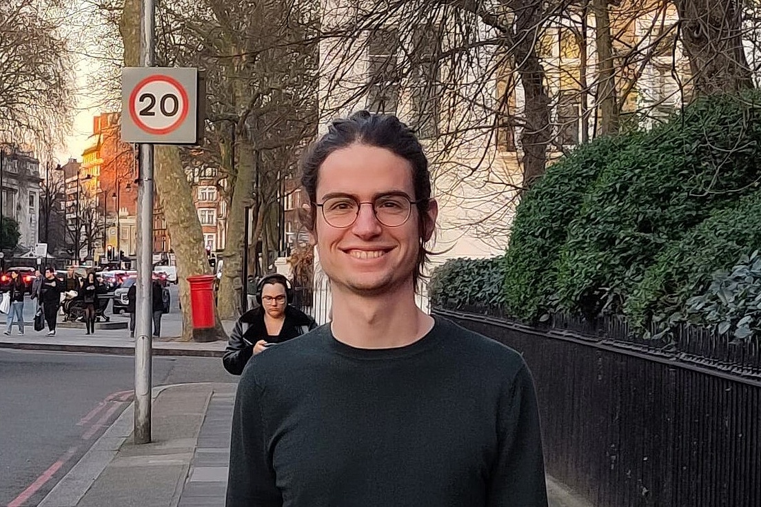 Junger Mann mit langem Haar und Brille lächelt vor rotem Londoner Doppeldeckerbus, 20-km/h-Schild und roter Telefonzelle bei Sonnenuntergang.