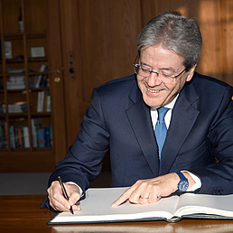 Paolo Gentiloni sitzt lächelnd an einem Tisch und schreibt in ein Buch.