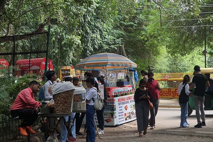 Lebhafter Straßenverkaufsstand in Delhi unter gestreiftem Sonnenschirm mit Snacks wie Pani Puri, umgeben von Menschen in Sommerkleidung, Bäumen, Coca-Cola-Kühler und einem gelben Tuk-Tuk.