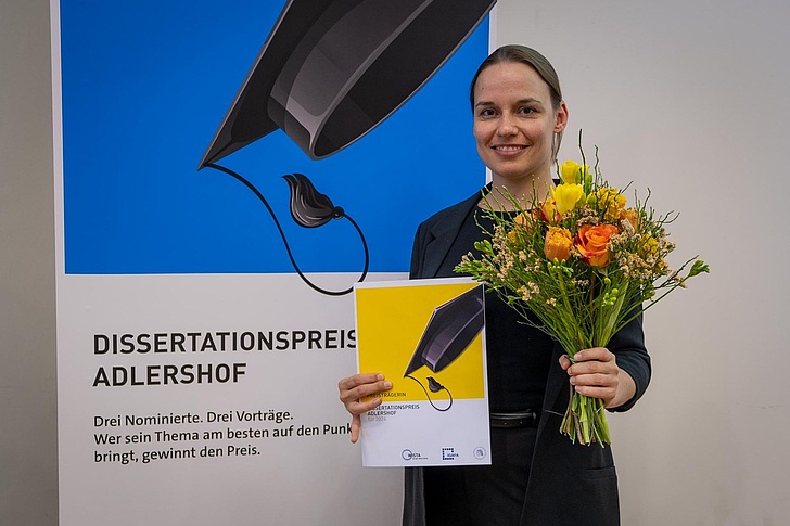 Dr. Kristin Klaue hält einen Blumenstrauß und ihre Urkunde in der Hand. Im Hintergrund ist ein Plakat des Dissertationspreises Adlershof zu sehen.
