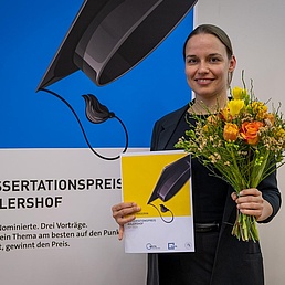 Dr Kristin Klaue holds a bouquet of flowers and her certificate. A poster for the Adlershof Dissertation Prize can be seen in the background.