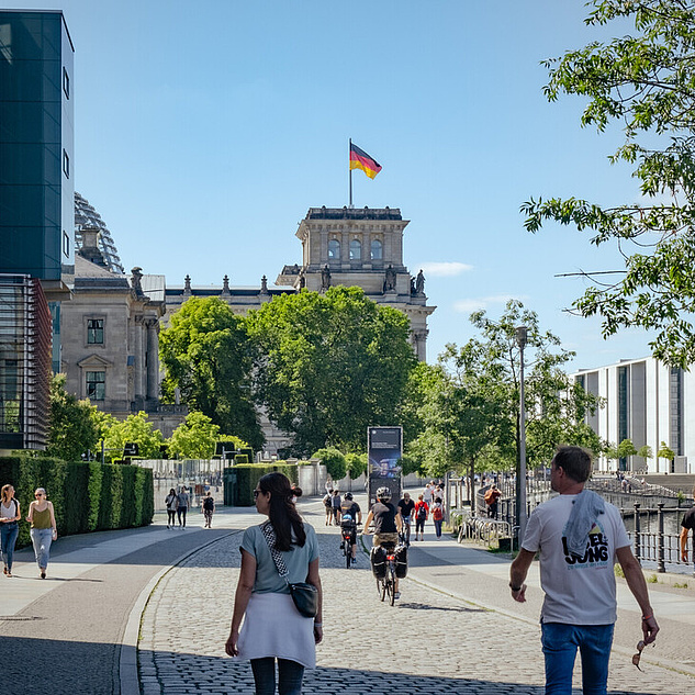Das Reichstagsgebäude in Berlin Das Bild zeigt eine belebte Straßenszene in Berlin. Im Hintergrund ist das Reichstagsgebäude mit der deutschen Flagge auf dem Dach zu sehen. Das Gebäude ist von grünen Bäumen umgeben, die einen Kontrast zum historischen Bauwerk bilden. In der Mitte des Bildes verläuft ein breiter Gehweg, auf dem Fußgänger*innen und Radfahrende unterwegs sind. Einige Personen scheinen sich zu unterhalten, während andere einfach nur spazieren gehen. Die Atmosphäre wirkt entspannt und lebendig.