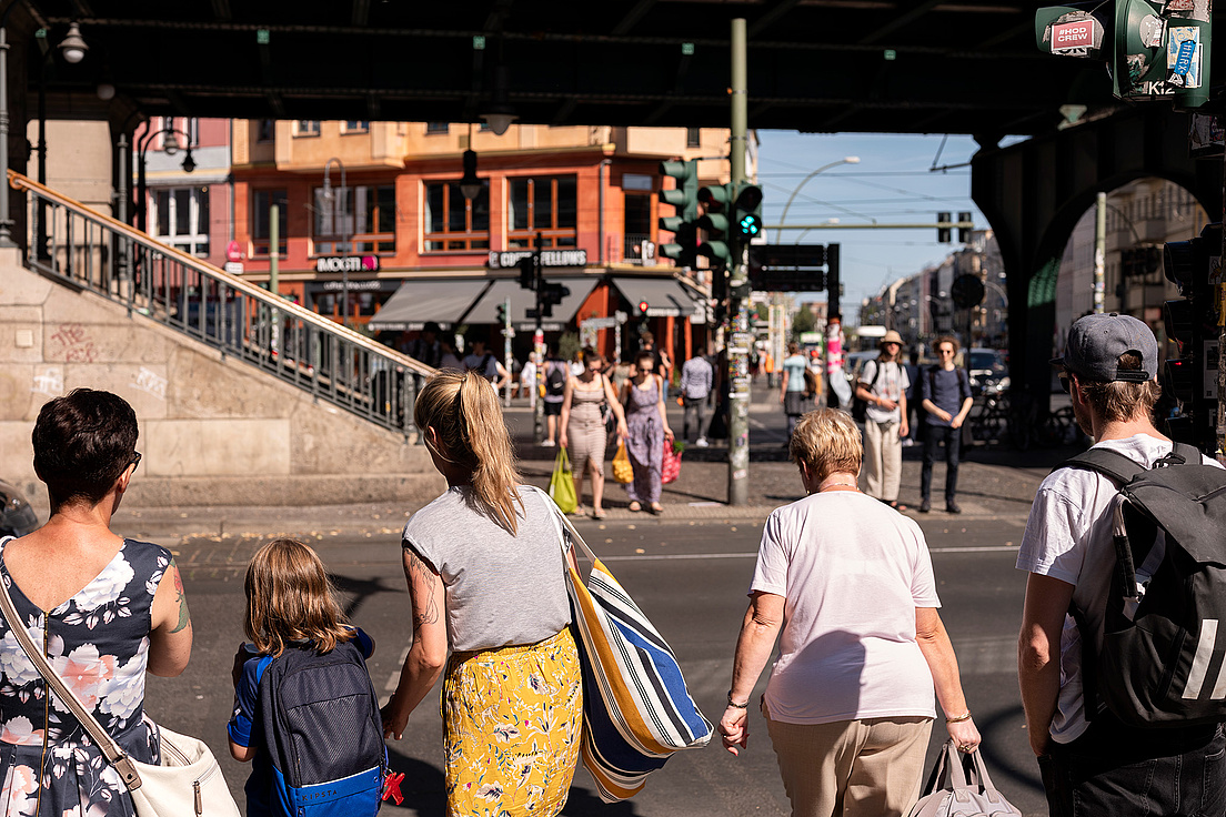 Das Bild zeigt eine belebte Straßenszene in einer Stadt. Mehrere Menschen sind zu sehen, die entweder auf der Straße stehen oder darauf warten, sie zu überqueren. Im Vordergrund befinden sich fünf Personen, darunter eine Frau mit einem Kind, das einen Rucksack trägt. Im Hintergrund sind weitere Fußgänger zu sehen, die sich auf der Straße bewegen. Es gibt auch einige Geschäfte und Cafés mit Schildern. 